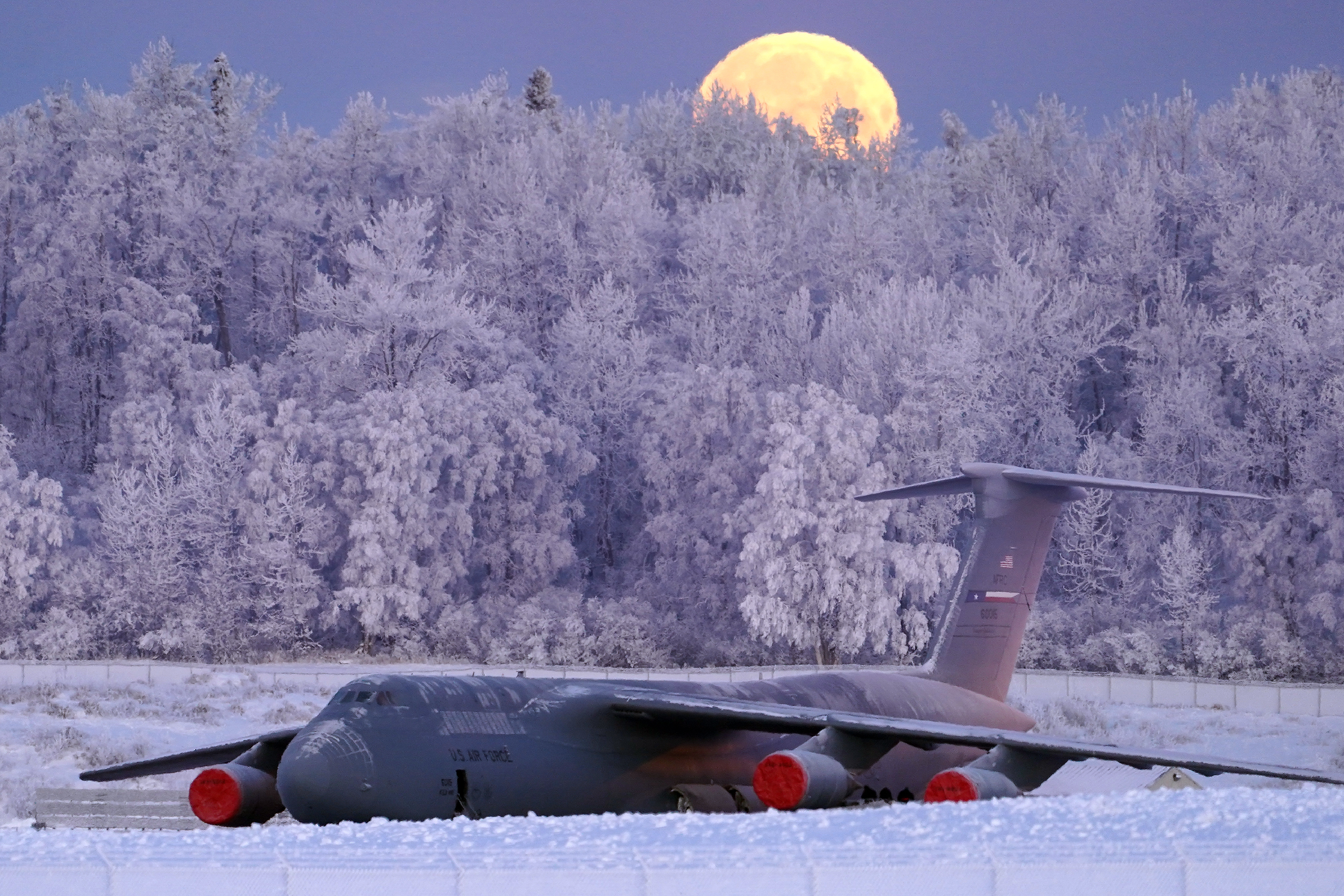 The waning gibbous moon is seen above a U.S. Air Force C-5 Galaxy transport aircraft on the flight line at Joint Base Elmendorf-Richardson, Alaska, Jan 10, 2020, as hoarfrost engulfs nearby trees in -16F weather while airmen work around the plane. (Justin Connaher/Air Force)