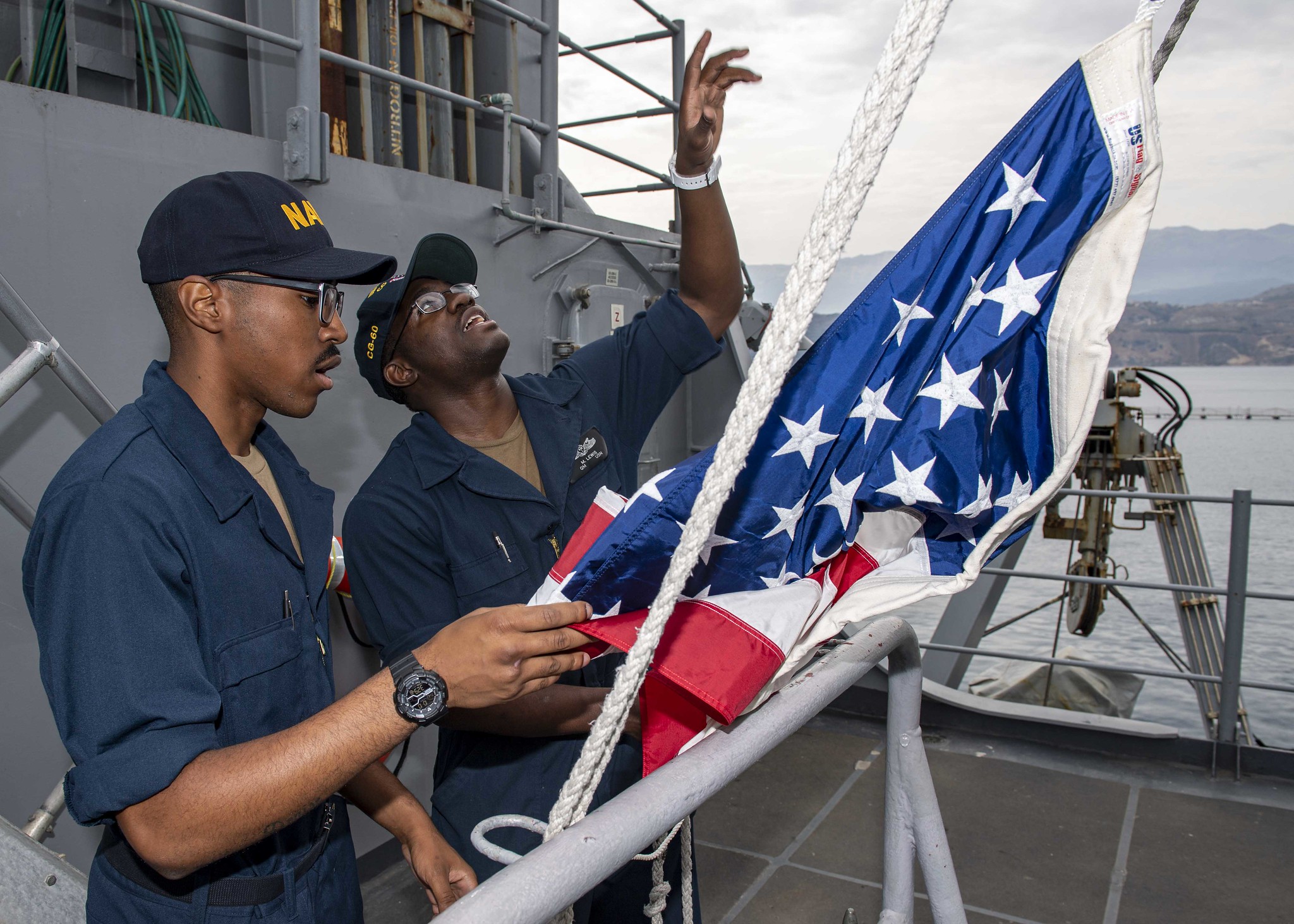 Quartermaster Seaman Marquis Lewis, right, instructs Quartermaster Seaman Khalil Hancock, on how to hoist the American flag as the Ticonderoga-class guided-missile cruiser USS Normandy (CG 60) prepares to depart Souda Bay, Greece, on Oct. 10, 2019. (Mass Communication Specialist 2nd Class Michael H. Lehman/Navy)