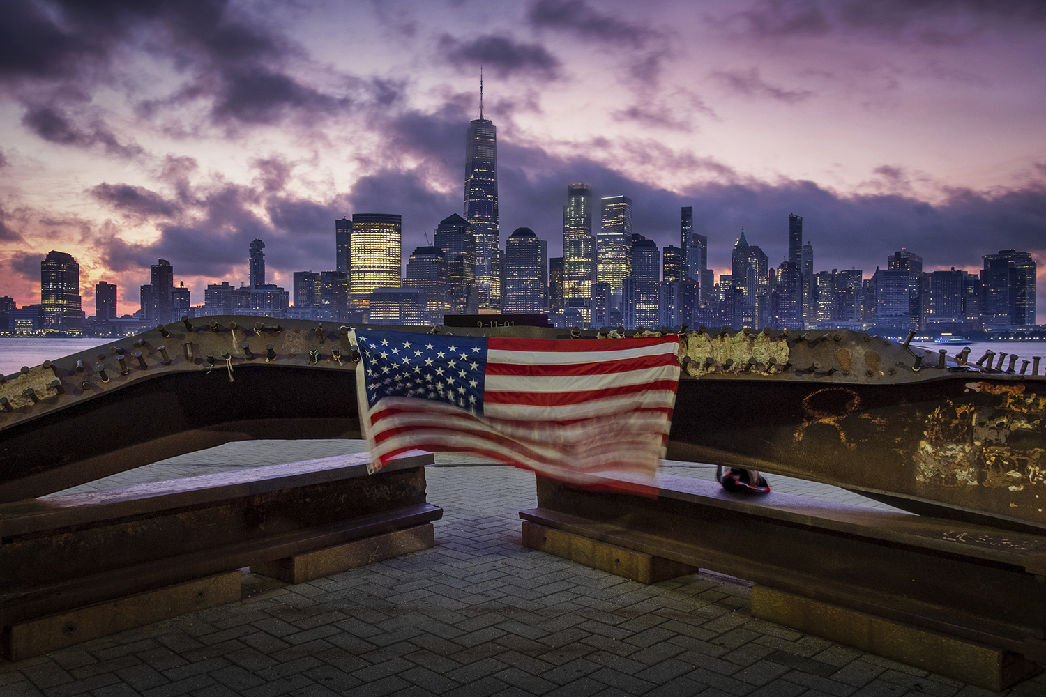 A U.S. flag hanging from a steel girder, damaged in the Sept. 11, 2001, attacks on the World Trade Center, blows in the breeze at a memorial in Jersey City, N.J., Sept. 11, 2019, as the sun rises behind One World Trade Center building and the re-developed area where the Twin Towers of World Trade Center once stood in New York City on the 18th anniversary of the attacks. (J. David Ake/AP)